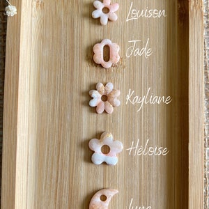 Puede incluir: Cinco amuletos de resina en forma de flor y media luna, de color rosa y blanco con destellos dorados. Los amuletos están dispuestos verticalmente sobre una superficie de madera. Los nombres Louison, Jade, Kayliane, Heloise y Luna están escritos junto a cada amuleto.