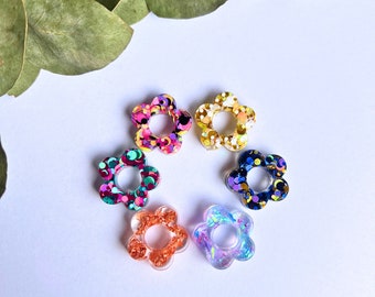 Könnte beinhalten: Sechs blütenförmige Harzverzierungen mit Glitzer. Die Blüten sind kreisförmig angeordnet, jede mit einem anderen Farbschema, darunter Rosa, Gold, Blau und schillernd. Der Hintergrund ist weiß mit einem Zweig grüner Blätter.