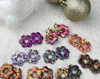 Könnte beinhalten: Sortiment an bunten Glitzer-Blumenohrringen. Die Ohrringe sind aus Harz und haben einen goldenen Reifen. Die Blumen sind in verschiedenen Farben, darunter Lila, Rosa, Orange und Grün.