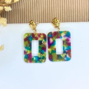 Pode incluir: Um par de brincos quadrados com uma parte superior de metal dourado e um corpo de resina translúcida marmorizada e colorida. A resina tem um esquema de cores verde, azul, roxo e rosa.