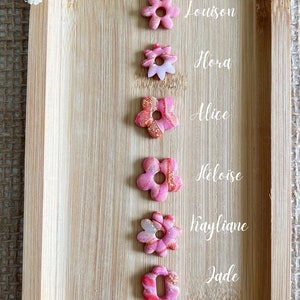 Può includere: Otto perline a forma di fiore rosa e bianco con dettagli dorati, disposte verticalmente su una superficie di legno. Ogni perlina ha un nome scritto accanto: Cassie, Louison, Flora, Alice, Héloïse, Kayliane, Jade e Ambre.