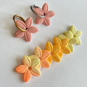 Peut inclure: Un ensemble de six boucles d'oreilles en fleurs en argile polymère dans des tons de rose, jaune et orange. Les boucles d'oreilles sont en dégradé, avec les fleurs roses en haut et les fleurs orange en bas. Les fleurs sont toutes des étoiles à cinq branches avec un trou au centre.