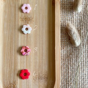 Può includere: Quattro perline di resina a forma di fiore in un vassoio di legno. Le perline sono rosa, bianche con strisce dorate, rosa con vortici dorati e rosse.