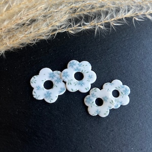 Op de afbeelding: Vier witte, bloemvormige knopen met een blauw bloemenpatroon en een gat in het midden. De knopen liggen op een zwart oppervlak.