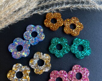 Könnte beinhalten: Sortiment von acht glitzernden Ohrringen in Blumenform in verschiedenen Farben, darunter Gold, Blau, Grün und Pink. Die Ohrringe werden auf einer schwarzen Oberfläche präsentiert.