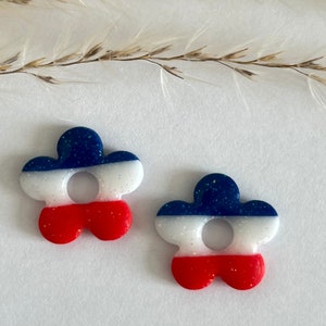 Peut inclure: Une paire de boucles d'oreilles en forme de fleur rouge, blanche et bleue avec des paillettes. Les boucles d'oreilles sont en résine et ont une finition pailletée.