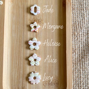 Può includere: Sette perline di ceramica bianca a forma di fiore con strisce dorate e rosa. Le perline sono etichettate con i nomi Flora, Jade, Morgane, Heloise, Alice, Lucy e Kayliane.