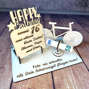 Könnte beinhalten: Ein Kuchenaufsatz aus Holz zum Geburtstag mit den Worten "HAPPY Birthday sweet 16" und anderem deutschen Text. Es zeigt ein Fahrrad und einen aufgerollten Euro-Schein. Die Basis ist weiß.