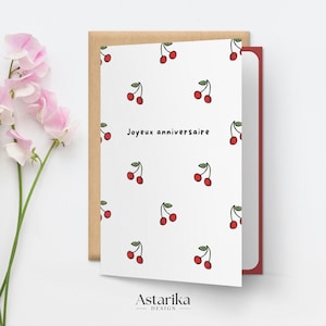 May include: A white card with a red border and a pattern of red cherries. The card says "Joyeux anniversaire" in black text.