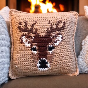May include: A brown crocheted pillow with a deer face design. The deer's antlers, eyes, and nose are crocheted in different colors.