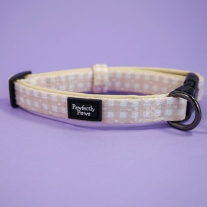 Könnte beinhalten: Ein beigefarbenes Hundehalsband mit einem weißen Karomuster und einer schwarzen Schnalle. Das Halsband hat ein kleines rechteckiges Schild mit der Aufschrift "Pawfectly Paws".