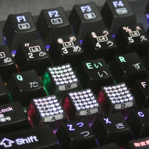 May include: Close-up of a black mechanical keyboard with illuminated keys. The keys feature English letters, numbers, symbols, and Japanese characters. Some keys are embellished with small, sparkling accents.