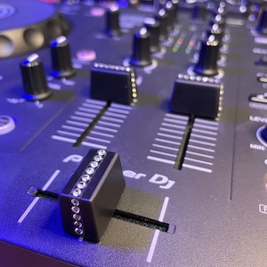 May include: A black DJ mixer with a silver knob and a black fader with a row of rhinestones. The mixer has a white label that says "Pioneer DJ".