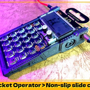 Puede incluir: Un sampler de música Pocket Operator negro y gris con una funda protectora. La funda tiene una superficie texturizada y un diseño antideslizante. El sampler tiene una pequeña pantalla y una cuadrícula de botones.