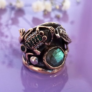 Könnte beinhalten: Ein einzigartiger Ring mit einem Frosch-Design, verziert mit grünen und klaren Edelsteinen. Der Ring hat eine dunkle Metallbasis mit goldfarbenen Akzenten und einem großen, irisierenden grünen Stein. Der Ring ist vor einem lila Hintergrund platziert.