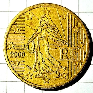 May include: A gold-coloured French coin from 2000. The coin depicts a woman with flowing hair and outstretched arms, encircled by stars. The letters "RF" are present, alongside the year "2000".