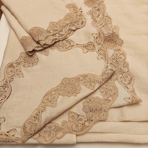Pode incluir: Uma toalha de mesa de linho creme com bordas de renda castanho-claro intrincadas. A renda apresenta padrões florais e geométricos, criando um design delicado e elegante. A toalha de mesa está dobrada, mostrando a guarnição de renda detalhada.