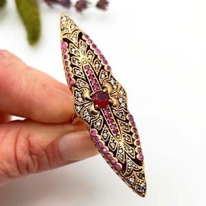 以下が含まれることがあります： 金色のステートメントリング。細長い装飾的なデザインで、中央に赤い宝石、その周りに小さな赤と透明な石、複雑なフィリグリーのディテールが施されています。白い背景に、手でリングが持たれています。