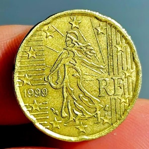May include: A gold-coloured coin with intricate detailing. The coin depicts a woman in flowing robes, encircled by stars, with the year 1999 clearly visible. The word "RE" is also present. The coin is held between fingers.