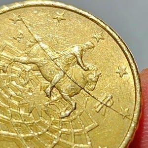 May include: Close-up of a gold coin with a detailed relief of a person riding a bull, surrounded by stars and geometric patterns. The coin has a shiny surface and is held by a person's fingers.