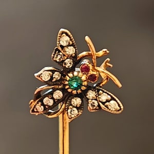 Op de afbeelding: Een goudkleurige bloembroche met een centrale smaragdgroene steen en rode en heldere edelstenen. De broche heeft een speldsluiting en staat tegen een donkergrijze achtergrond. Waarschijnlijk een vintage of antiek stuk.