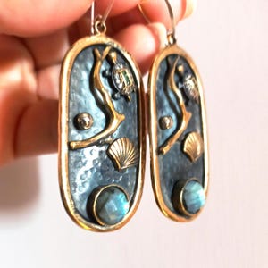 Puede incluir: Un par de pendientes de plata y oro con textura martillada. Los pendientes presentan una rama con una tortuga, una concha y una piedra preciosa azul.