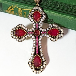 May include: A large ornate gold cross pendant with red and white gemstones. The cross is decorated with a border of small white gemstones and has a red gemstone at the centre.