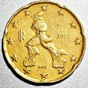 May include: A gold-coloured coin with a distinctive design. The coin shows a central figure with outstretched arms, a stylized depiction of a person, and the year 2002. The coin's edge is decorated with stars.