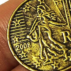 May include: Close-up of a gold-coloured coin with an intricate design. The coin shows a woman and the year 2008. The coin has a worn, antique appearance, with visible stars and text around the edges.