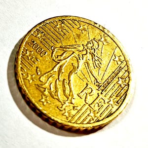 Puede incluir: Una moneda de euro de 2000 de color dorado. La moneda presenta una mujer con cabello suelto y estrellas. La palabra "EURO" es visible. La moneda es redonda con un borde estriado.