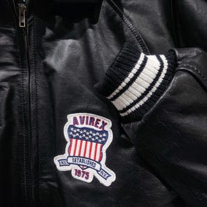 May include: Black leather jacket with a zipper closure and a ribbed knit cuff with white stripes. The jacket features a patch with the text "AVIREX ESTABLISHED 1975 N.Y." in red, white, and blue.