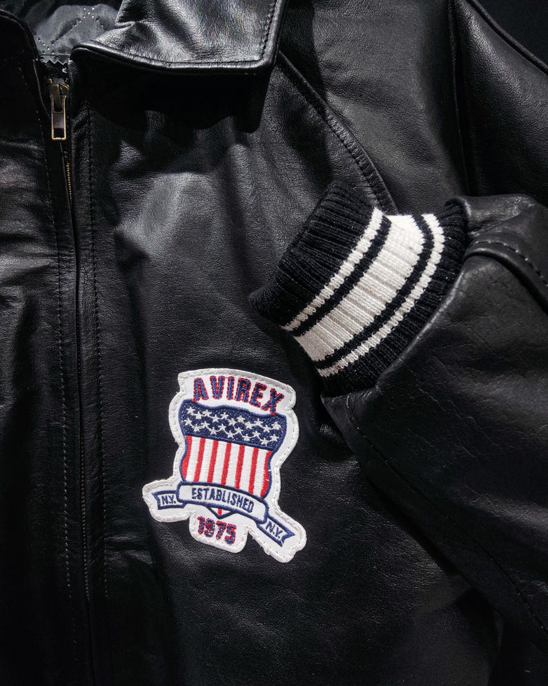 May include: Black leather jacket with a zipper closure and a ribbed knit cuff with white stripes. The jacket features a patch with the text "AVIREX ESTABLISHED 1975 N.Y." in red, white, and blue.