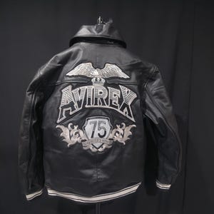 Puede incluir: Chaqueta de cuero negro con un &aacute;guila bordada en blanco y plata y el logotipo "AVIREX" en la espalda. La chaqueta presenta un parche "75" y un dobladillo a rayas blancas y plateadas. Una chaqueta bomber cl&aacute;sica.