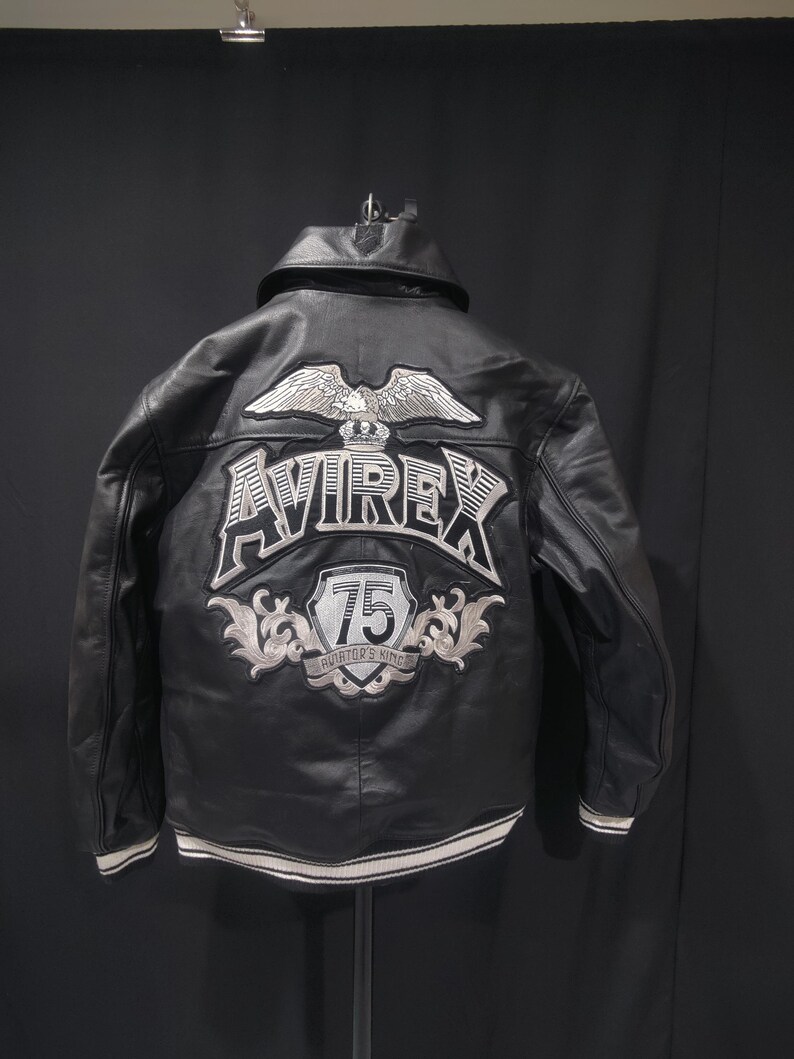 Puede incluir: Chaqueta de cuero negro con un &aacute;guila bordada en blanco y plata y el logotipo "AVIREX" en la espalda. La chaqueta presenta un parche "75" y un dobladillo a rayas blancas y plateadas. Una chaqueta bomber cl&aacute;sica.
