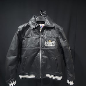 Puede incluir: Chaqueta de cuero negro con cremallera, cuello y pu&ntilde;os y dobladillo a rayas blancas. La chaqueta presenta el logo "AVIREX" en el pecho. Un estilo bombardero cl&aacute;sico.