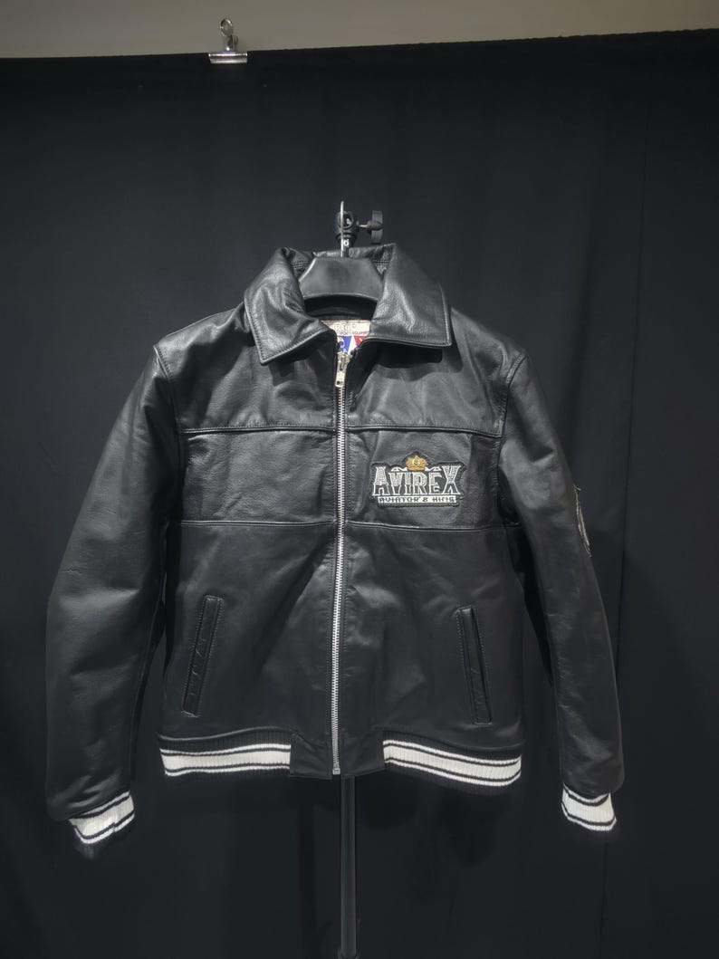 Puede incluir: Chaqueta de cuero negro con cremallera, cuello y pu&ntilde;os y dobladillo a rayas blancas. La chaqueta presenta el logo "AVIREX" en el pecho. Un estilo bombardero cl&aacute;sico.