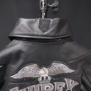 Puede incluir: Chaqueta de cuero negro con cuello y un emblema de &aacute;guila bordado. La chaqueta presenta el nombre de la marca "AVIREX" en una fuente audaz y estilizada. La chaqueta se muestra sobre un fondo oscuro.