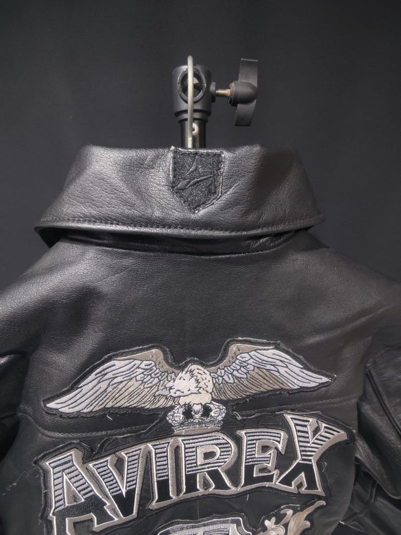 Puede incluir: Chaqueta de cuero negro con cuello y un emblema de &aacute;guila bordado. La chaqueta presenta el nombre de la marca "AVIREX" en una fuente audaz y estilizada. La chaqueta se muestra sobre un fondo oscuro.