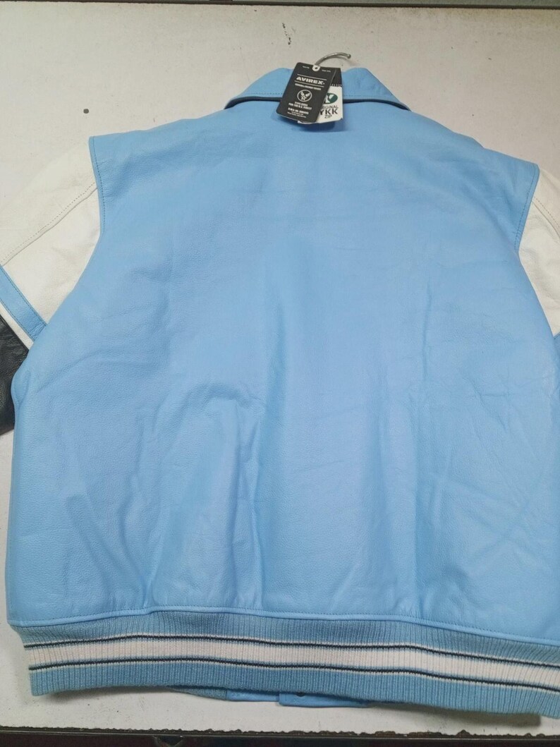 May include: Light blue varsity jacket with white sleeves and a striped hem. The jacket has a collar and a tag attached. The sleeves are white with a black stripe. The jacket is made of leather.