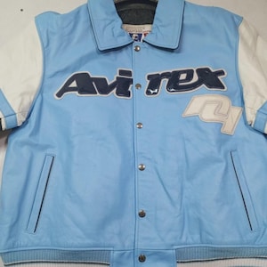 May include: Light blue leather jacket with white sleeves and the "Avirex" logo in navy blue and white. Features a button-up front, pockets, and a ribbed hem with white stripes. A classic varsity style.