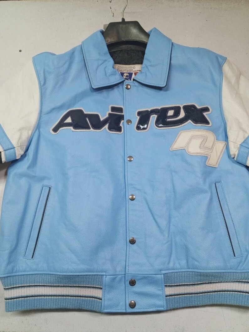 May include: Light blue leather jacket with white sleeves and the "Avirex" logo in navy blue and white. Features a button-up front, pockets, and a ribbed hem with white stripes. A classic varsity style.