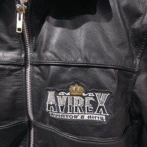 Puede incluir: Chaqueta de cuero negro con cremallera plateada y cuello. La chaqueta presenta un parche con el texto "AVIREX AVIATOR & KING" y una corona dorada.