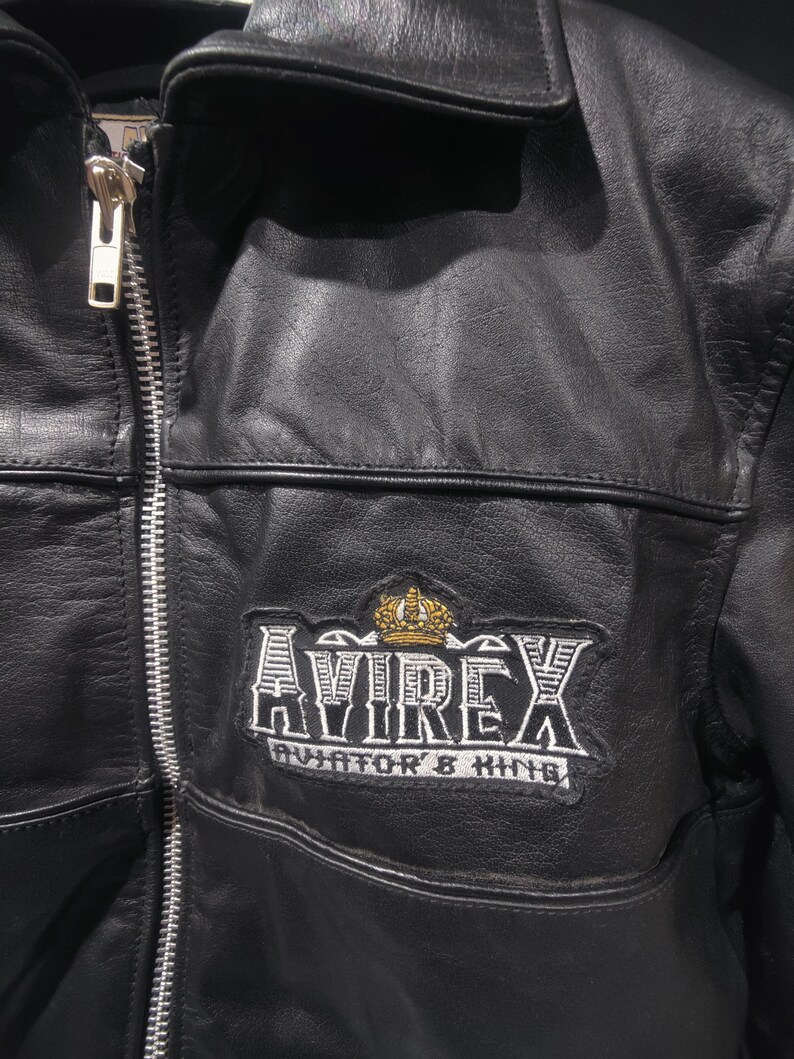Puede incluir: Chaqueta de cuero negro con cremallera plateada y cuello. La chaqueta presenta un parche con el texto "AVIREX AVIATOR & KING" y una corona dorada.