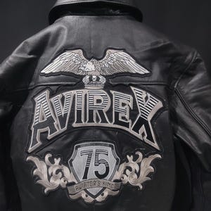 Puede incluir: Chaqueta de cuero negra con la palabra "AVIREX" y un parche de &aacute;guila en la espalda. La chaqueta tambi&eacute;n presenta un parche "75 Aviator's King". La chaqueta tiene cuello y forro negro.