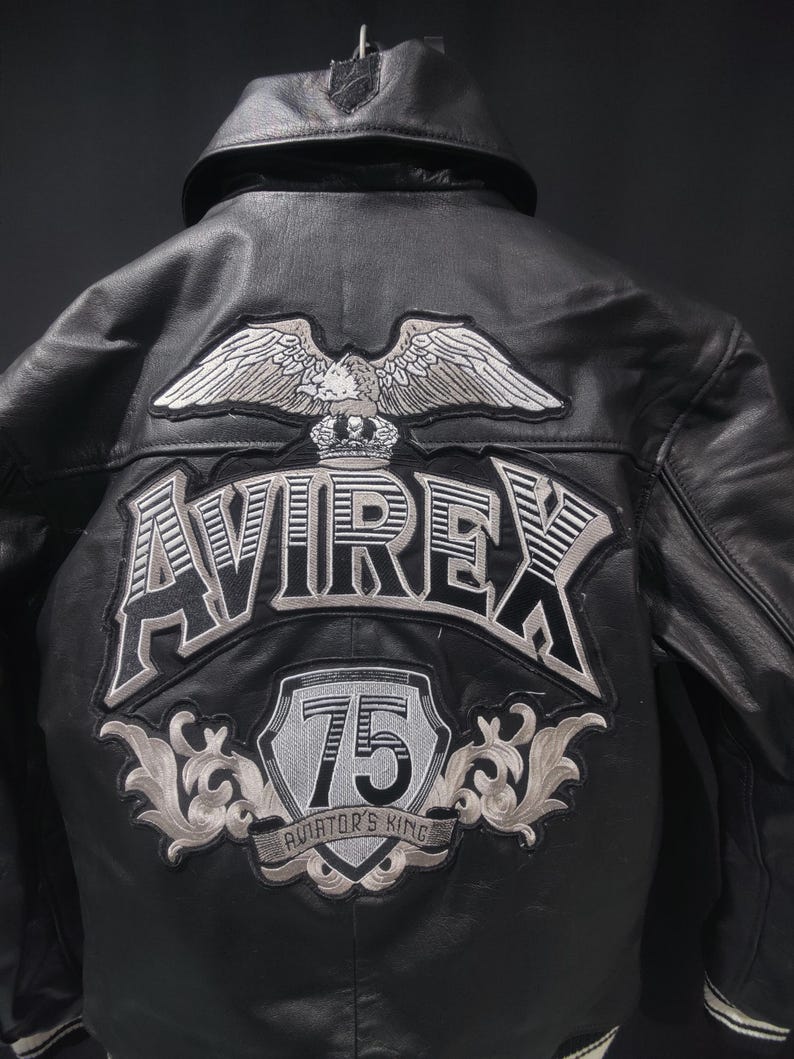Puede incluir: Chaqueta de cuero negra con la palabra "AVIREX" y un parche de &aacute;guila en la espalda. La chaqueta tambi&eacute;n presenta un parche "75 Aviator's King". La chaqueta tiene cuello y forro negro.