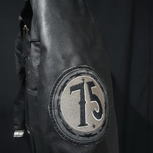 Puede incluir: Chaqueta de cuero negro con un parche circular en la manga con el n&uacute;mero 75 en gris y negro. La chaqueta tiene un dise&ntilde;o cl&aacute;sico con cuello y costuras visibles.