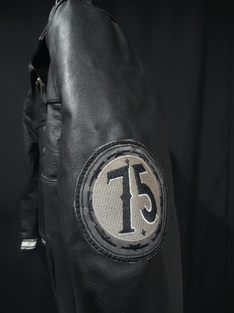 Puede incluir: Chaqueta de cuero negro con un parche circular en la manga con el n&uacute;mero 75 en gris y negro. La chaqueta tiene un dise&ntilde;o cl&aacute;sico con cuello y costuras visibles.