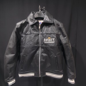 Puede incluir: Chaqueta de cuero negra con cremallera, cuello y pu&ntilde;os y dobladillo a rayas blancas y negras. La chaqueta presenta el logotipo "AVIREX" en el pecho. Un estilo bombardero cl&aacute;sico.