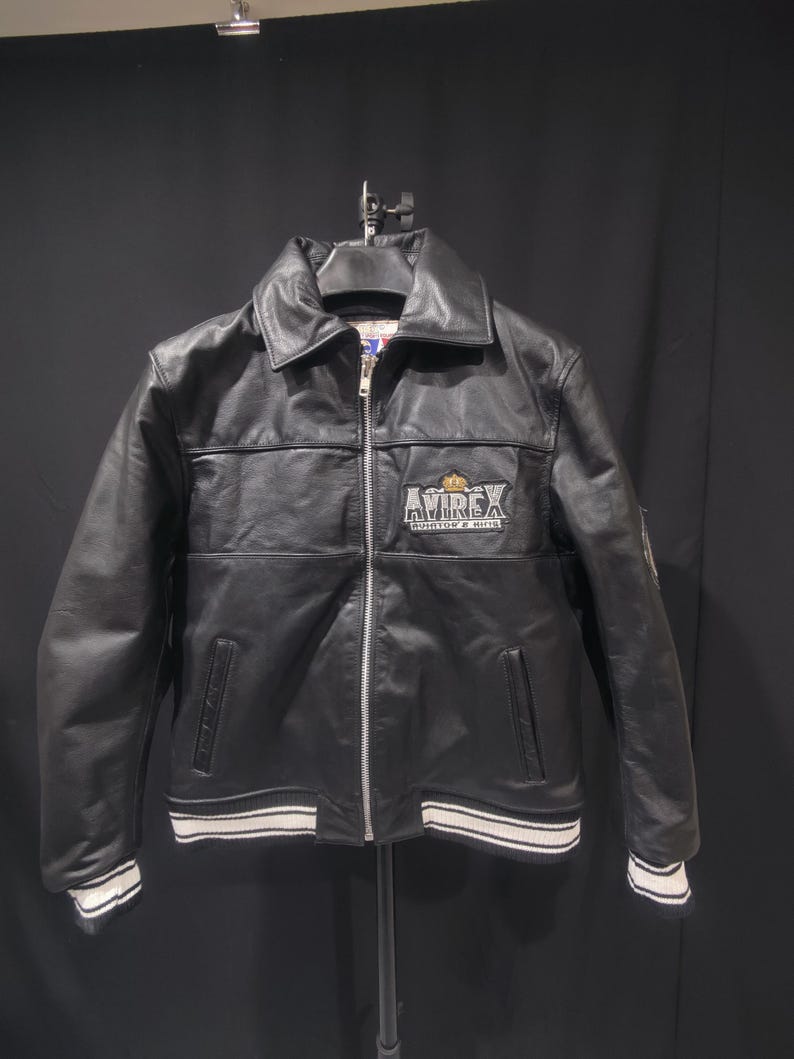 Puede incluir: Chaqueta de cuero negra con cremallera, cuello y pu&ntilde;os y dobladillo a rayas blancas y negras. La chaqueta presenta el logotipo "AVIREX" en el pecho. Un estilo bombardero cl&aacute;sico.