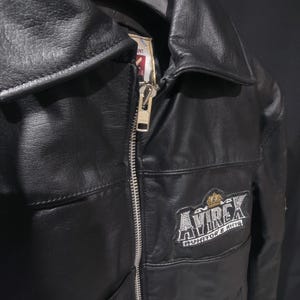 Puede incluir: Chaqueta de cuero negro con cremallera plateada y cuello con solapas. La chaqueta presenta el logotipo bordado "AVIREX" en el pecho. La chaqueta tiene un dise&ntilde;o cl&aacute;sico.